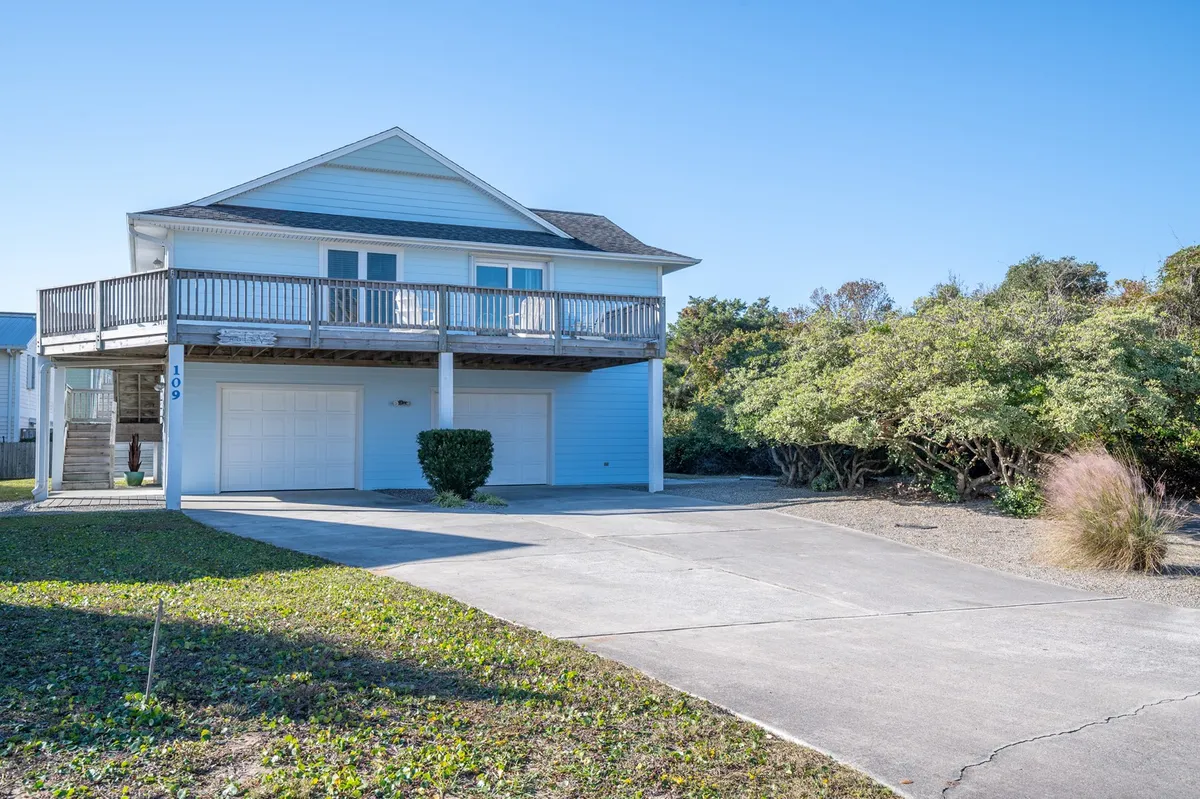 The exterior of the house is featured, showing the raised cottage design, balcony for sunset views, landscaped yard, and a spacious driveway perfect for gatherings or children playing. Family members can be seen relaxing on the balcony, hinting at the peaceful, private retreat atmosphere.