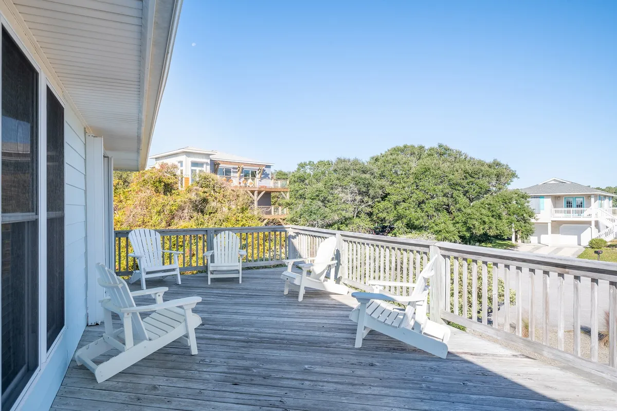 The image showcases the inviting exterior of a single-family beach house, featuring a spacious wooden deck adorned with classic Adirondack chairs. Lush greenery frames the space, creating a tranquil atmosphere perfect for relaxing, socializing, and taking in the refreshing coastal air. The deck hints at memorable sunrise coffees and relaxing evenings under the stars, just steps from Kure Beach.