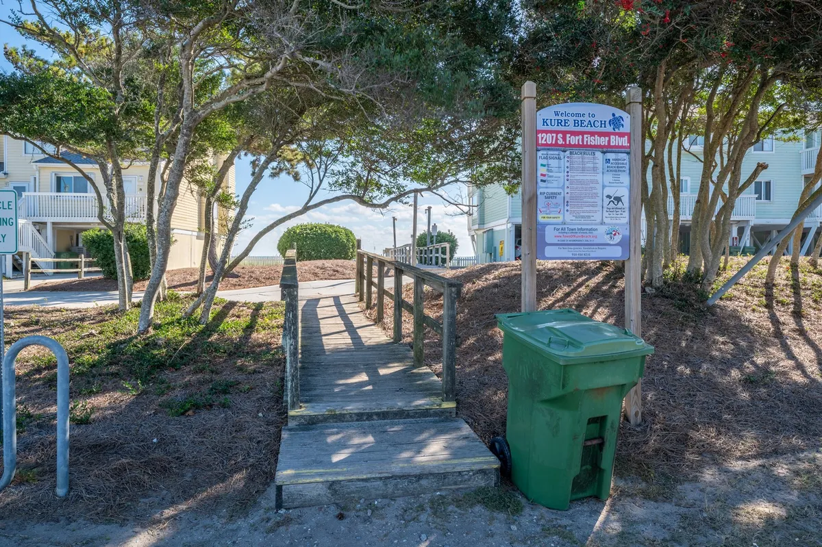 This view shows a wooden walkway with a beach access sign, surrounded by trees and greenery, leading toward the shoreline. The pathway suggests the anticipation and excitement of heading out for a day by the water, with space for families to walk together.