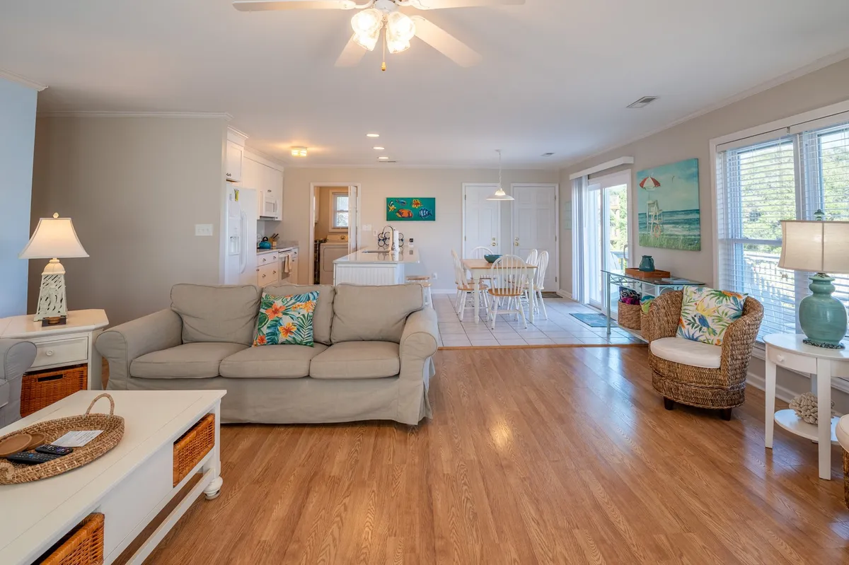 A warm and airy great room with a plush sofa, inviting dining table, vibrant artwork, and decorative light fixtures. Large windows let in ample sunlight, bathing the entire space in a golden glow and reinforcing the open, welcoming atmosphere ideal for family gatherings and meals.