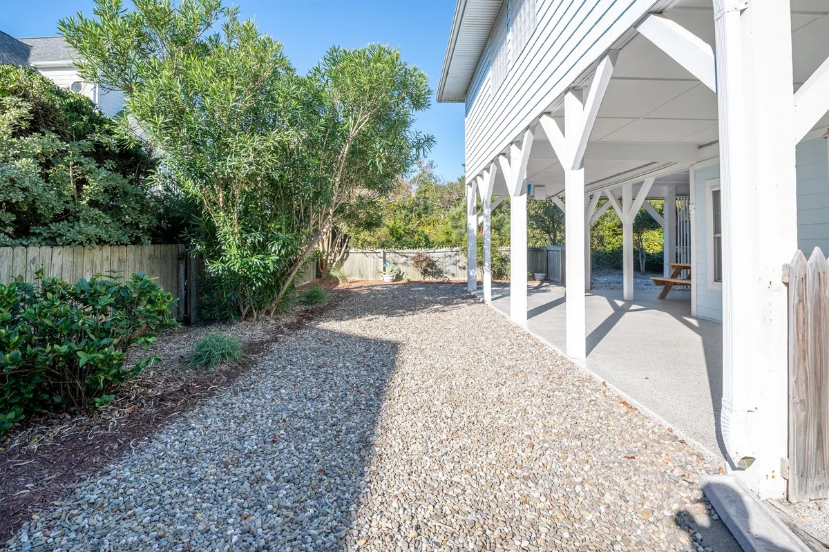 A tranquil exterior view of a gravel pathway bordered by lush greenery, leading to the shaded wraparound porch supported by sturdy beams. The porch is clearly a central feature, evoking images of relaxing mornings with coffee or unwinding in the evening, all while enjoying the fresh air and likely ocean views just beyond the frame.