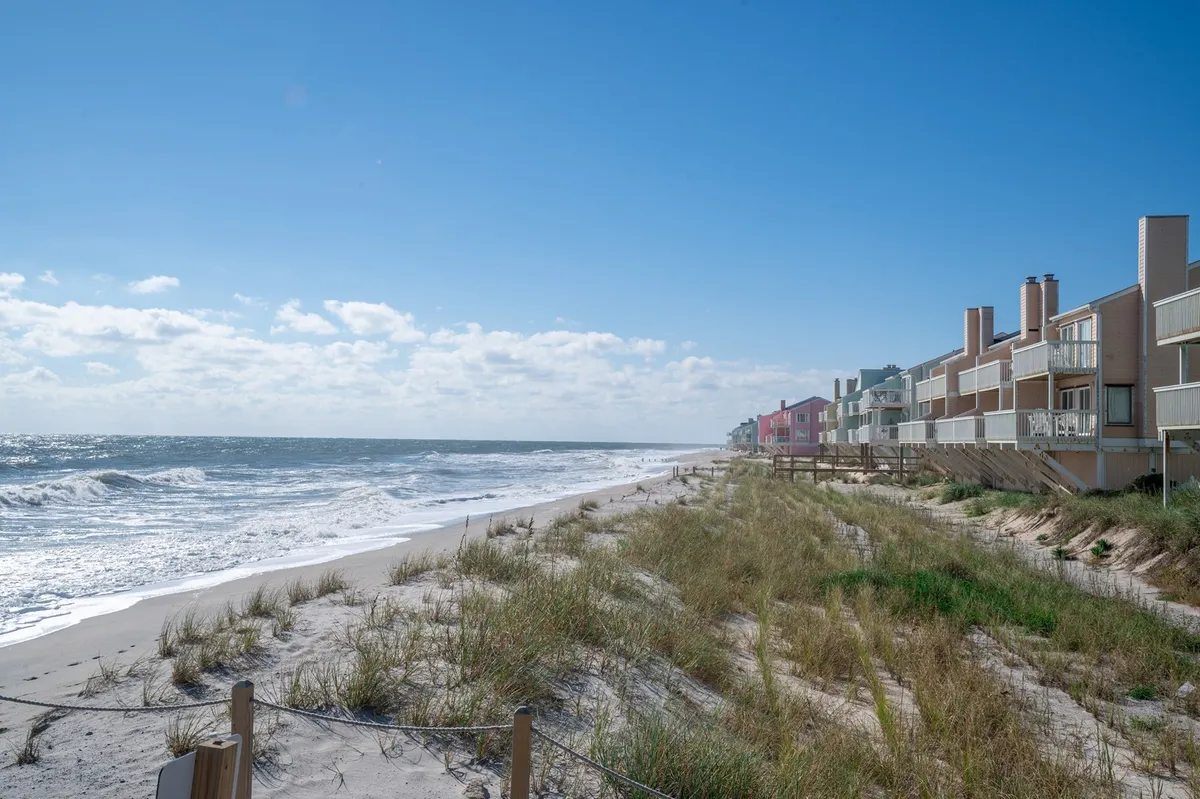 A wide view of the coastline, featuring rolling ocean waves, beach grass, and colorful beach houses, captures the local environment and the kinds of landscapes awaiting guests just minutes away. The scene is vibrant and lively, aligning with the adventure and discovery theme.