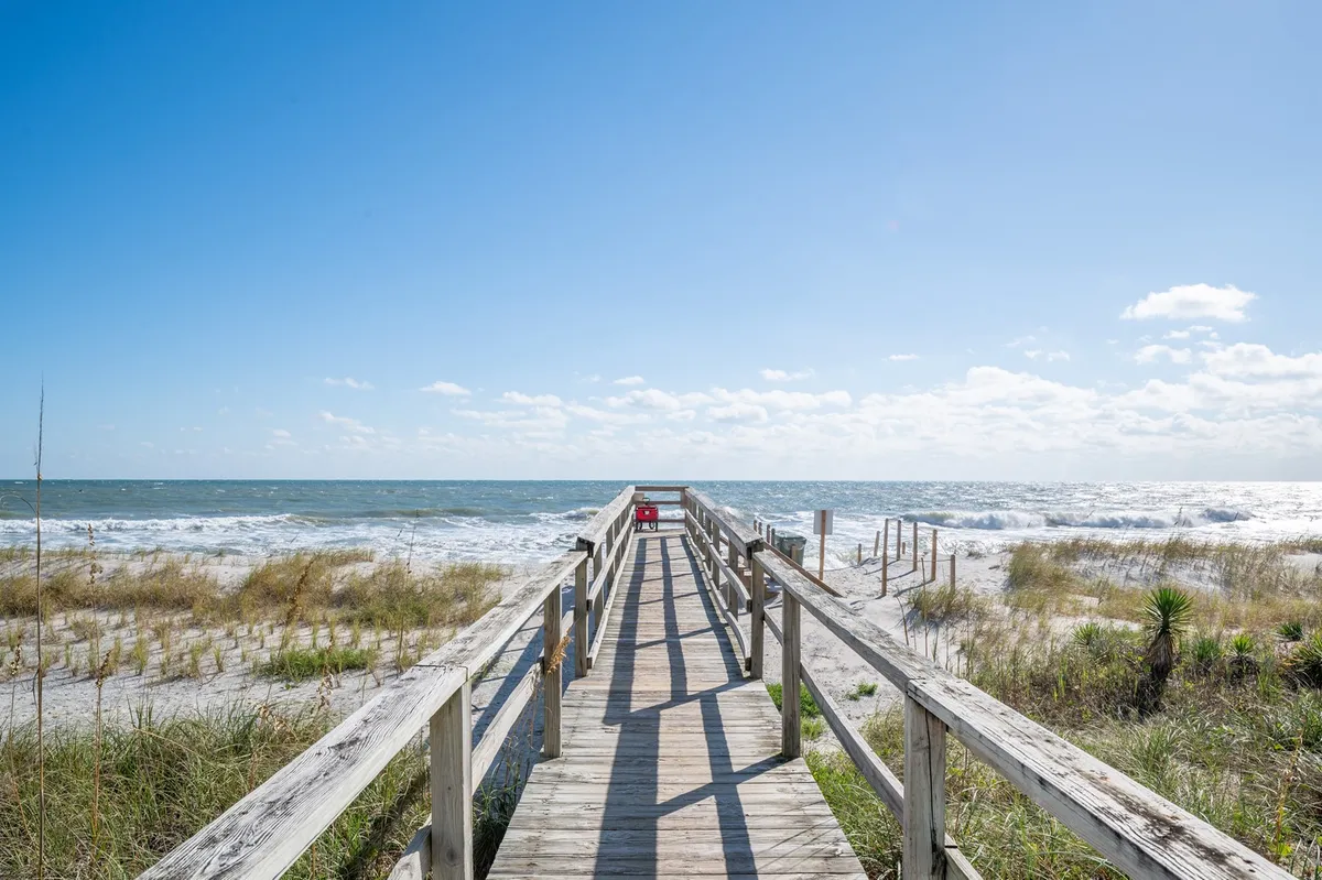 This image captures a serene wooden walkway leading over sand dunes toward the ocean under a sky filled with gentle clouds. The scene includes the inviting shoreline, emphasizing easy access to the beach and the natural beauty awaiting just steps from the cottage.