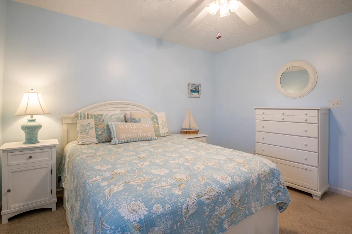 A tranquil bedroom featuring a bed with a light blue quilt, a nightstand with a lamp, a white dresser, and a decorative mirror. Natural light streams in, creating a peaceful and inviting atmosphere ideal for rest and relaxation.