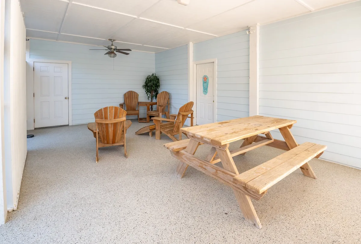 An inviting covered patio area featuring a wooden picnic table, Adirondack chairs, a potted plant, and a ceiling fan. The space is designed for gatherings and alfresco dining, offering a cozy yet airy ambience.