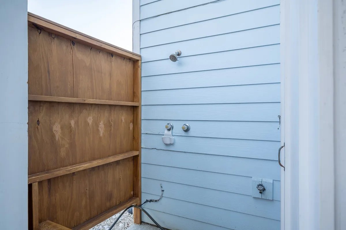 A refreshing outdoor shower enclosed by a wooden privacy fence, with blue siding visible in the background. The shower area looks clean, well-maintained, and provides privacy for rinsing off after beach outings.