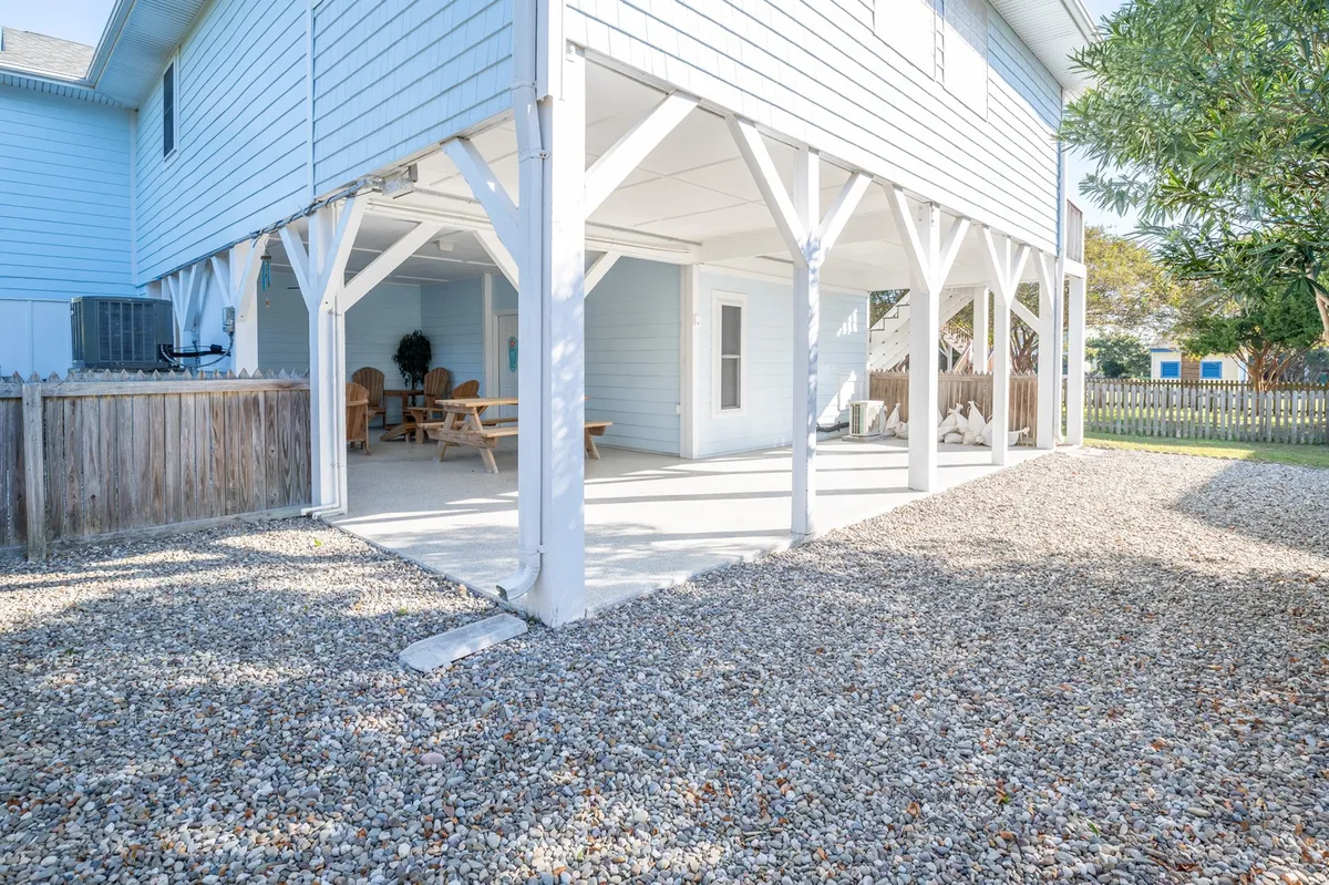 This exterior image showcases a relaxed outdoor seating area set on a gravel surface, framed by supportive columns. The space is clearly designed for social gatherings, with ample room for dining or simply lounging. The view hints at sunset skies, inviting shared meals or games in a laid-back setting. There’s a visible sense of openness and flexibility, ideal for everything from grilling to enjoying the ocean breeze.