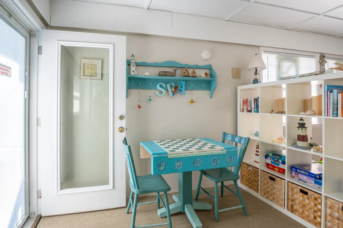 A cozy nook centered around a checkerboard table with blue chairs, complemented by a shelf with decorative items. The area is bright and inviting, encouraging game nights, reading, and simple relaxation.